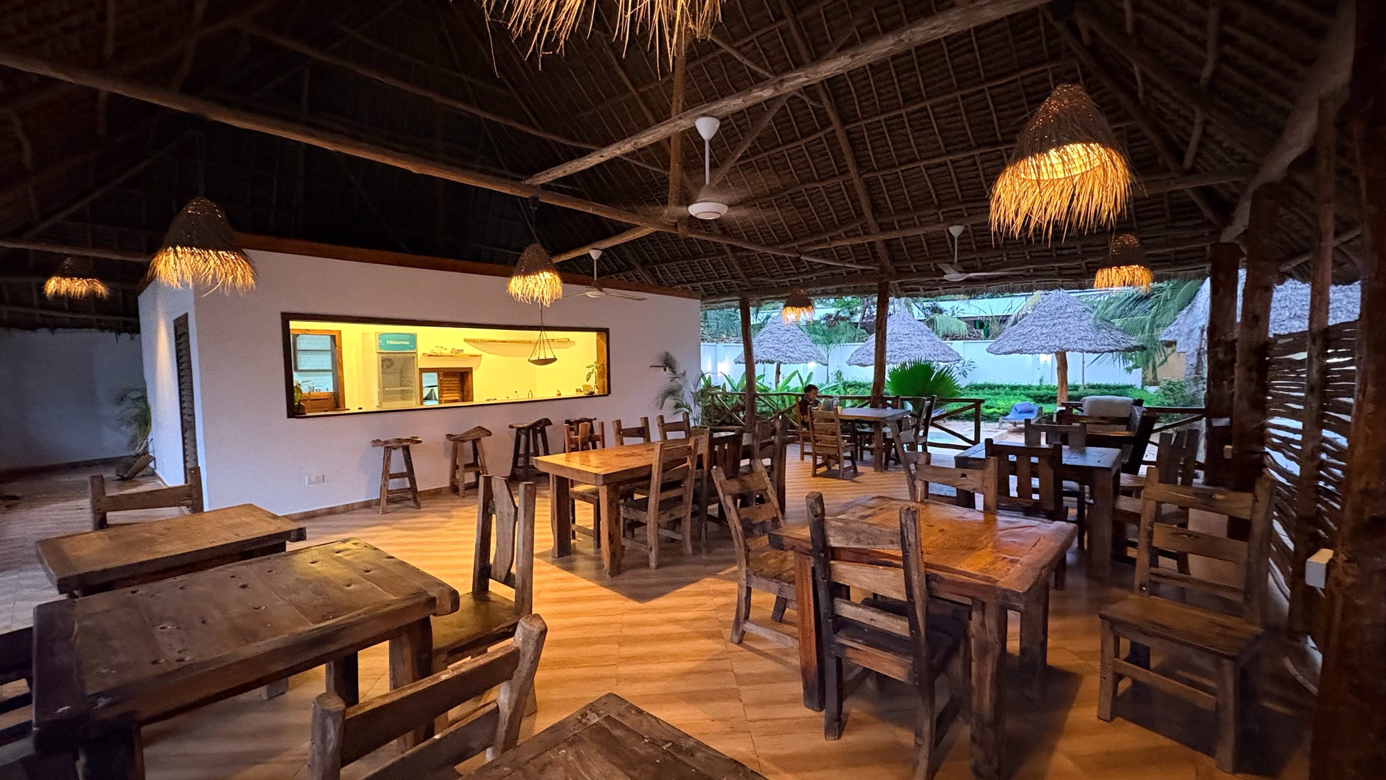 The airy on-site restaurant offers rustic wooden tables and chairs beneath a soaring thatched roof, opening onto the green garden beyond.
