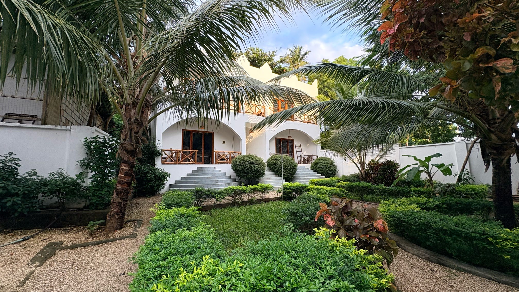 The whitewashed guest house rises above a lush manicured garden of clipped hedges and towering coconut palms, with curved wooden balconies and a stone staircase leading to the entrances.
