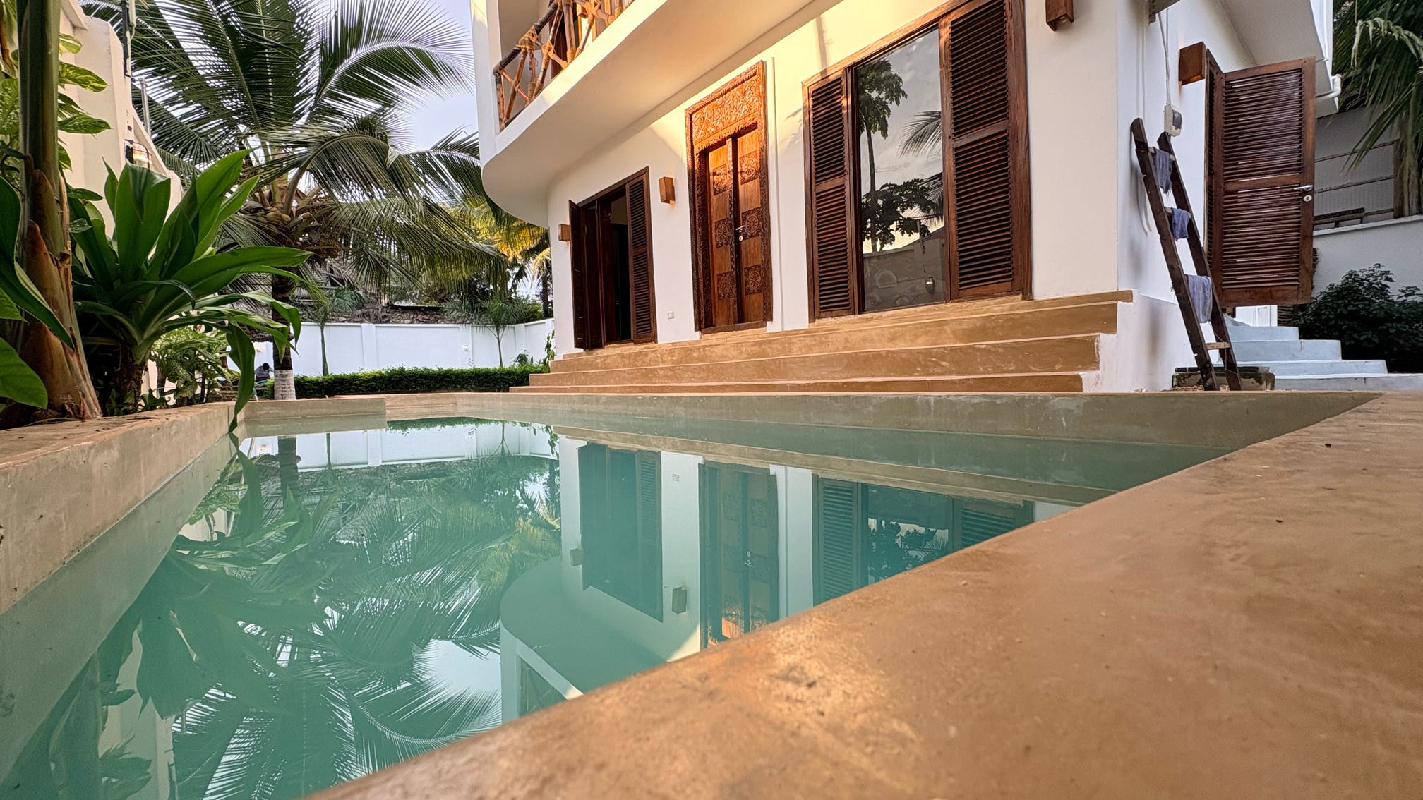 A serene swimming pool with glassy turquoise water mirroring the guest house, palm trees and bright blue sky.