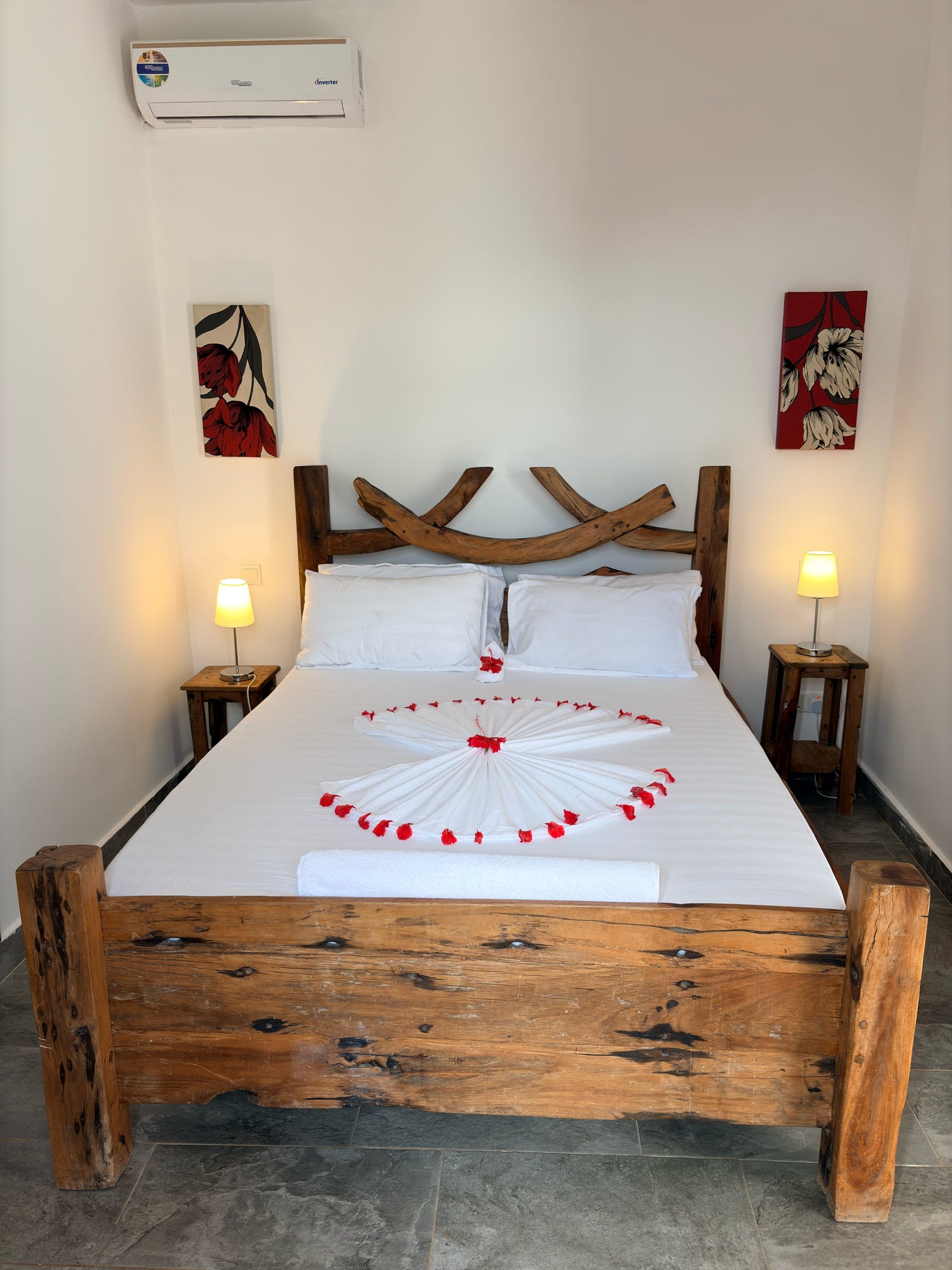 A neatly styled double room featuring a rustic wooden bed, twin bedside lamps, and a heart of red rose petals on crisp white linens.